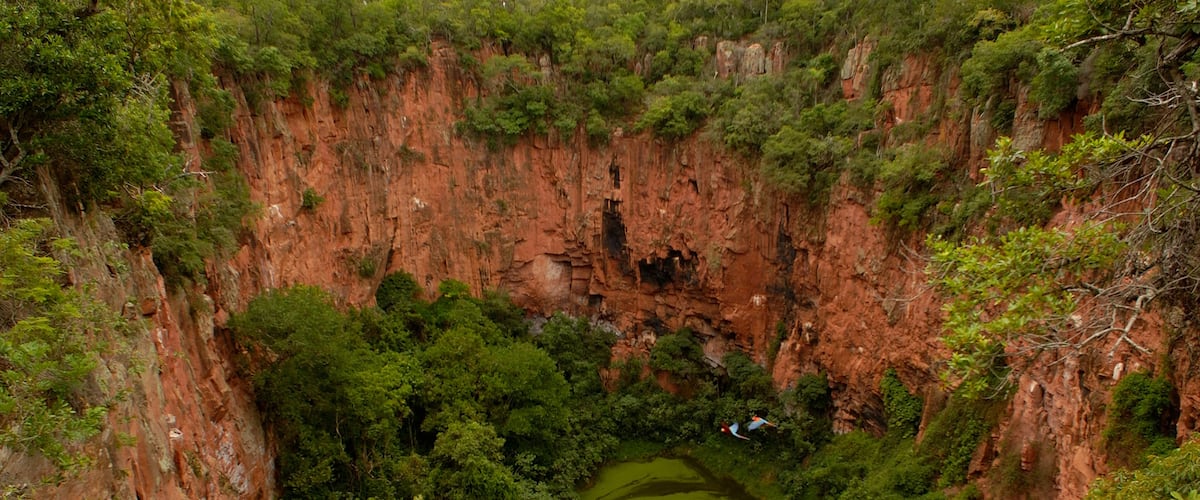 A deep canyon with a green pool at its base, surrounded by dense vegetation and rocky cliffs.