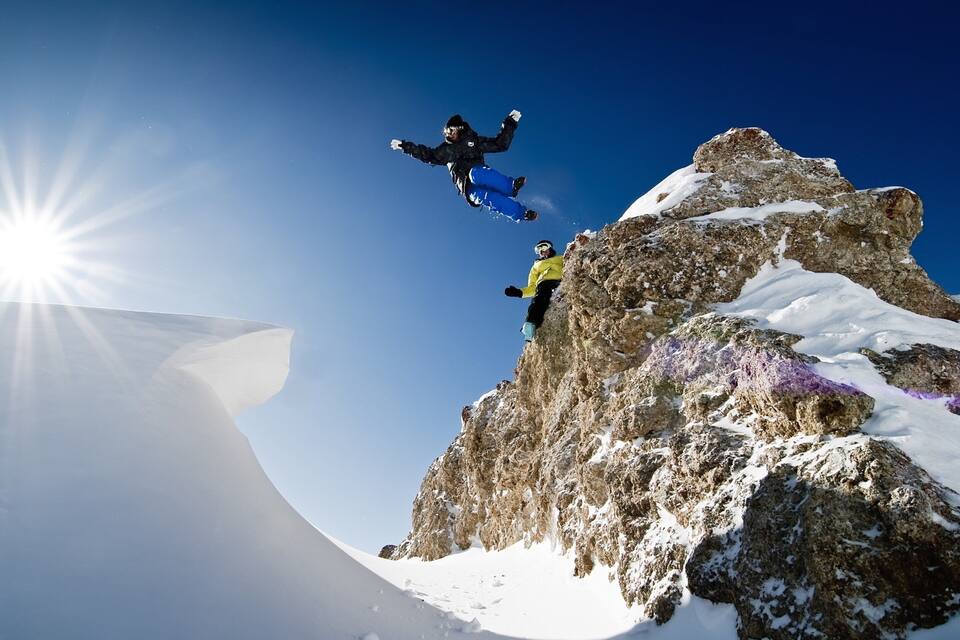 Un snowboardeur en plein vol au-dessus du sommet enneigé d’une montagne, avec le soleil qui brille en arrière-plan.