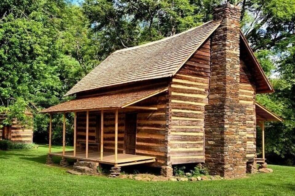 A rustic log cabin with a stone chimney, surrounded by lush green trees.