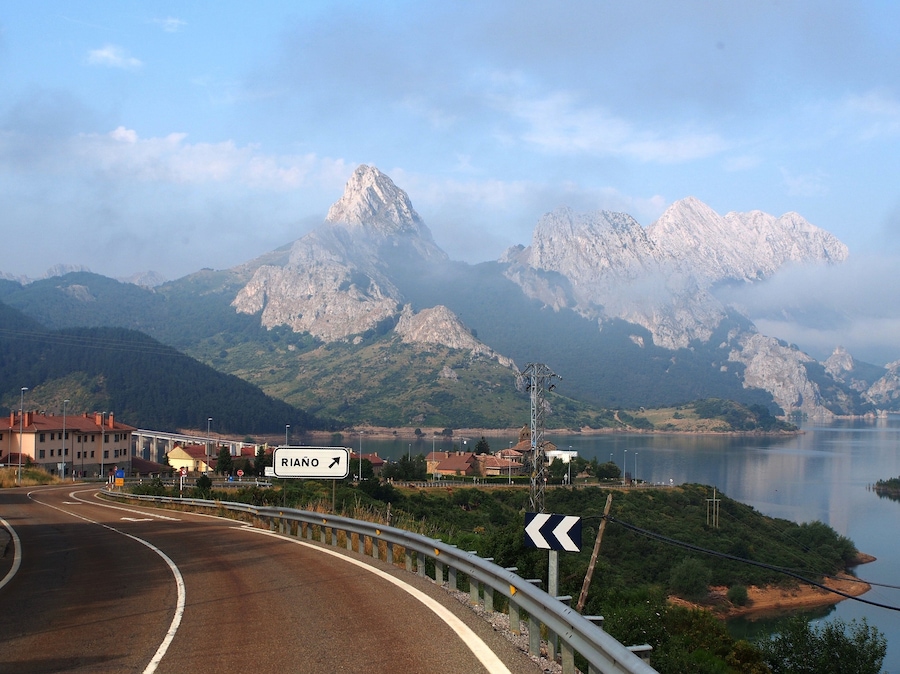 A winding road with a sign for RIAÑO, surrounded by mountains and a lake.