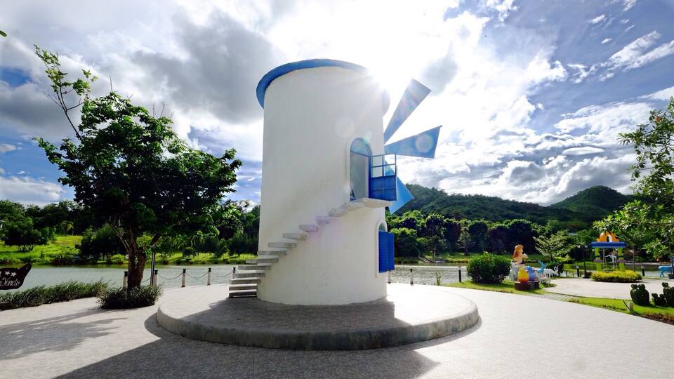 A windmill with a blue roof and a staircase leading up to it, surrounded by a park with trees and a lake.