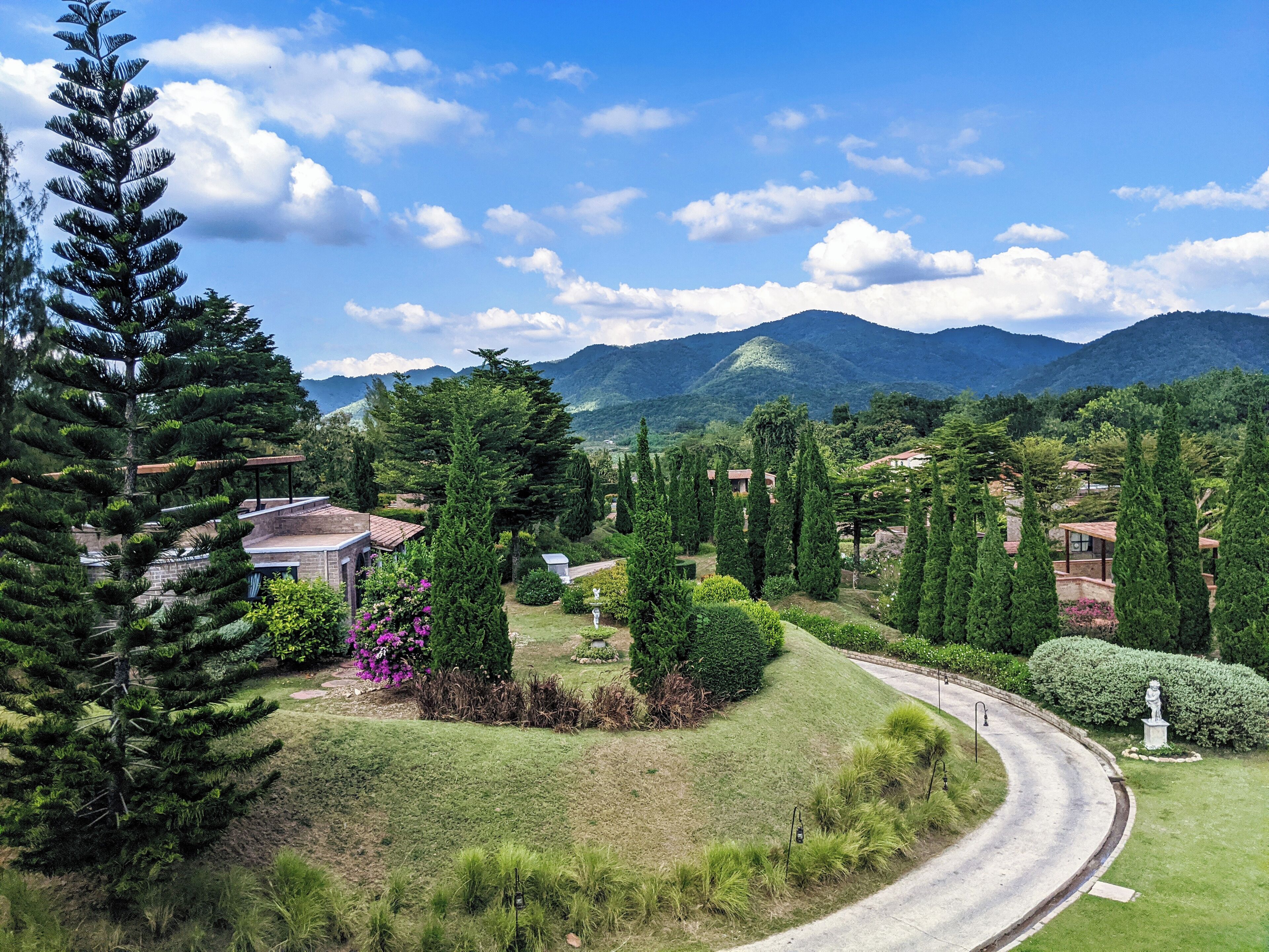 A winding road through a lush landscape with evergreen trees, a statue, and distant mountains under a blue sky.