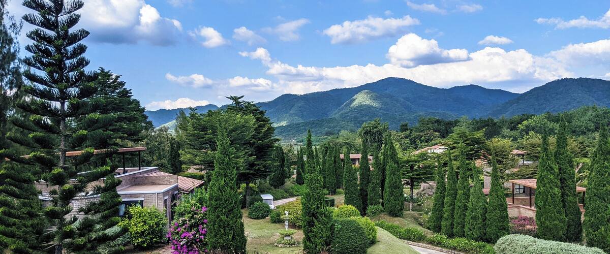 A winding road through a lush landscape with evergreen trees, a statue, and distant mountains under a blue sky.