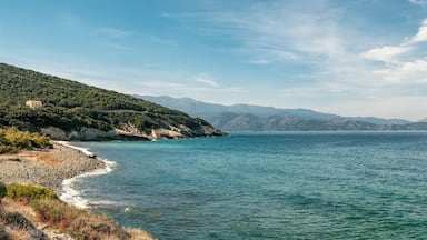 A coastal landscape with a rocky shoreline, a clear blue sea, and distant mountains under a partly cloudy sky.