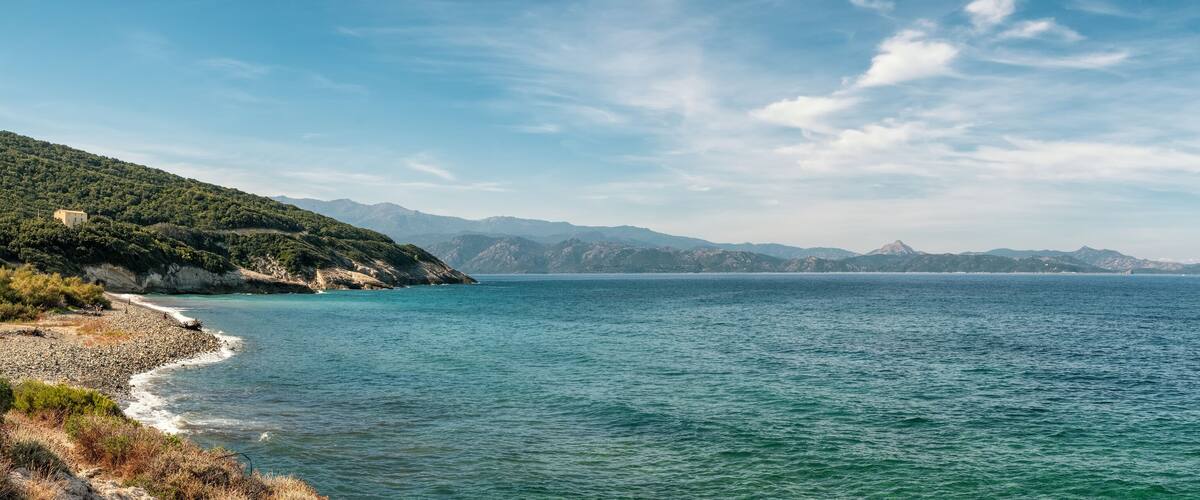 A coastal landscape with a rocky shoreline, a clear blue sea, and distant mountains under a partly cloudy sky.