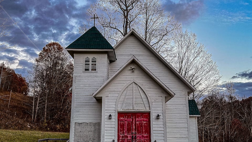 A small white church with a red door and a green roof, surrounded by trees and grass.