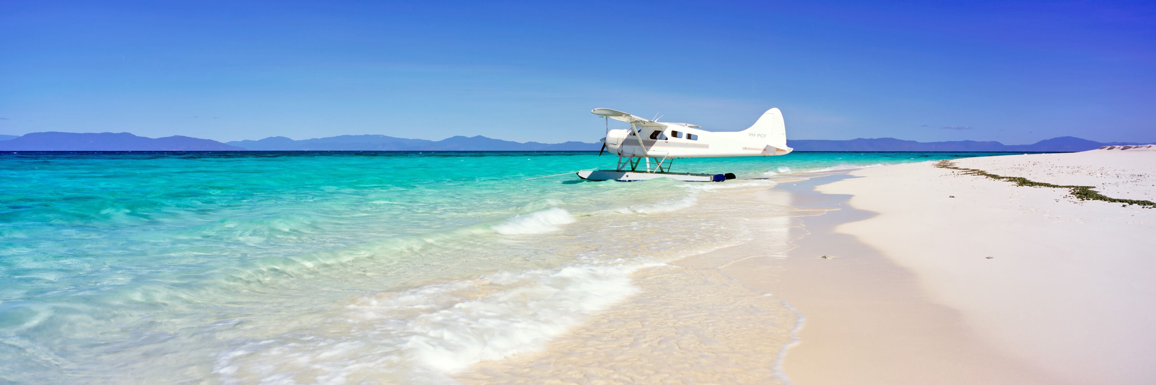 Un hidroavión blanco en una playa turquesa y cristalina, con montañas al fondo.