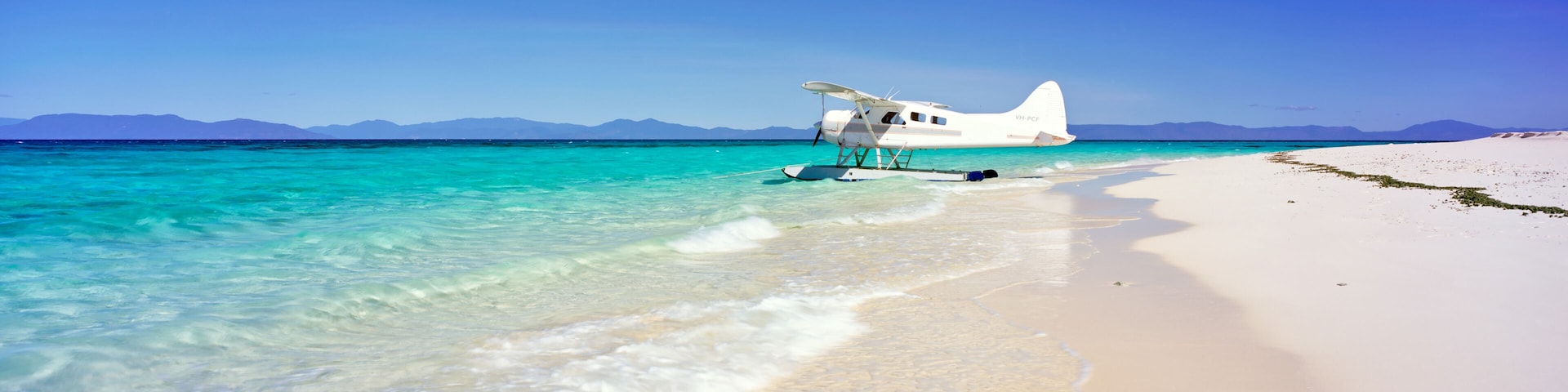 Un hydravion blanc sur une plage de sable turquoise, avec des montagnes en arriĂšre-plan.