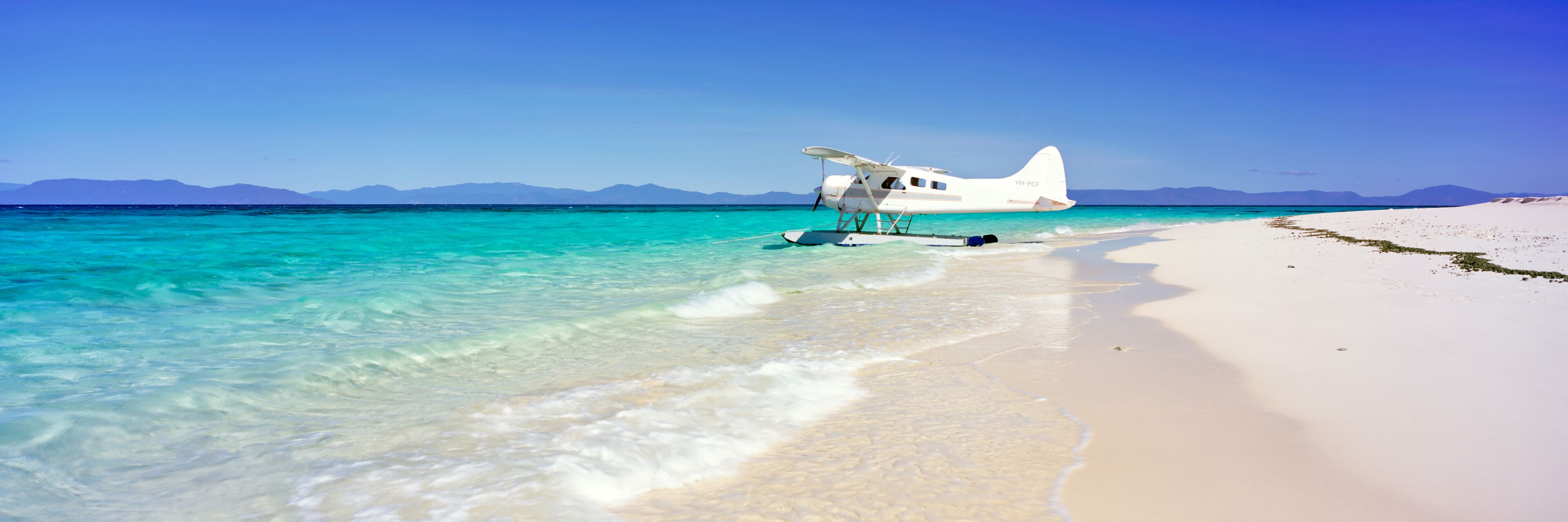 A white seaplane parked on a white sandy beach with clear turquoise water and mountains in the background.