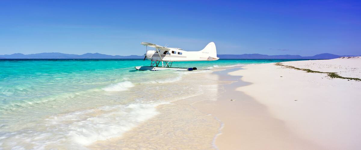 A white seaplane parked on a white sandy beach with clear turquoise water and mountains in the background.