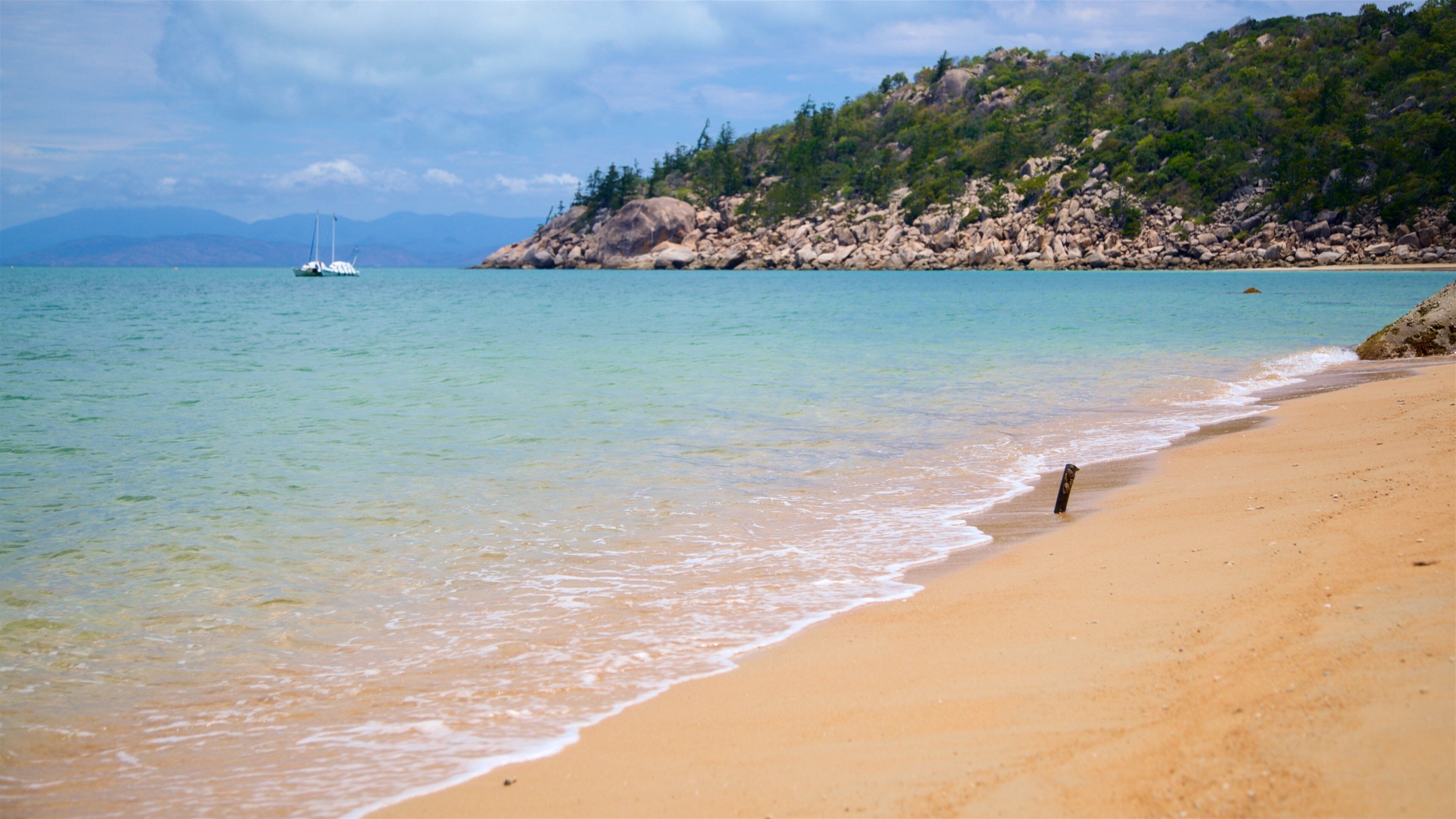 A sandy beach with gentle waves, a sailboat in the distance, and a rocky hillside with greenery.