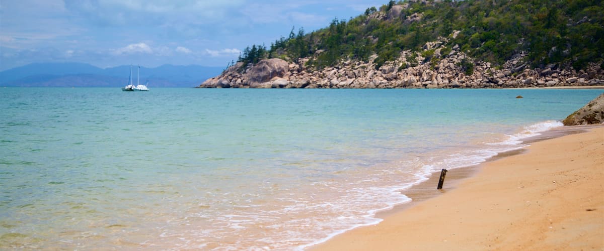A sandy beach with gentle waves, a sailboat in the distance, and a rocky hillside with greenery.