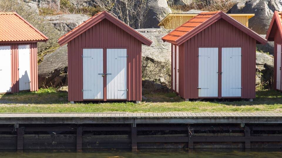 A row of red wooden boathouses by a calm body of water with orange buoys.