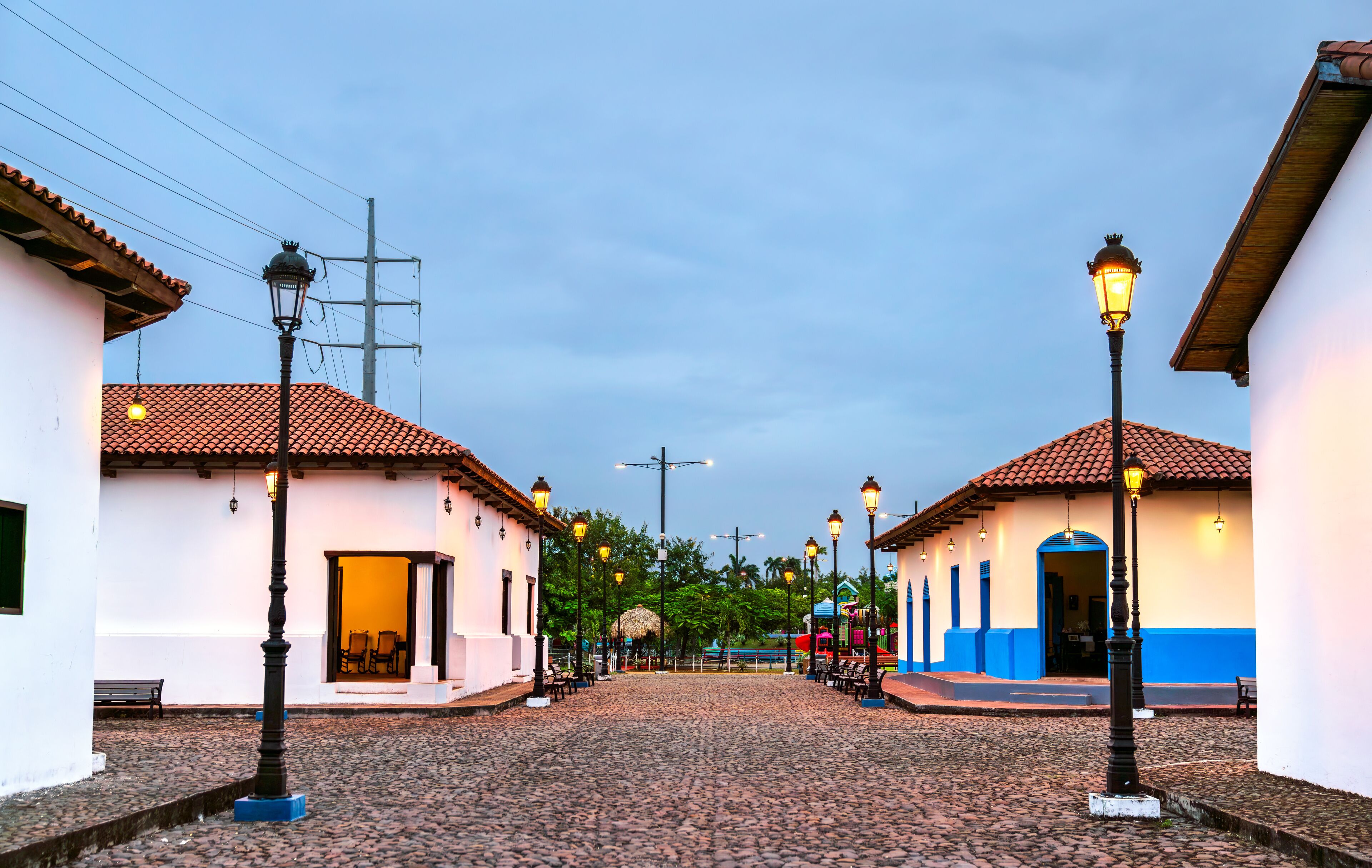 A cobblestone street with buildings, street lamps, and a thatched-roof structure.