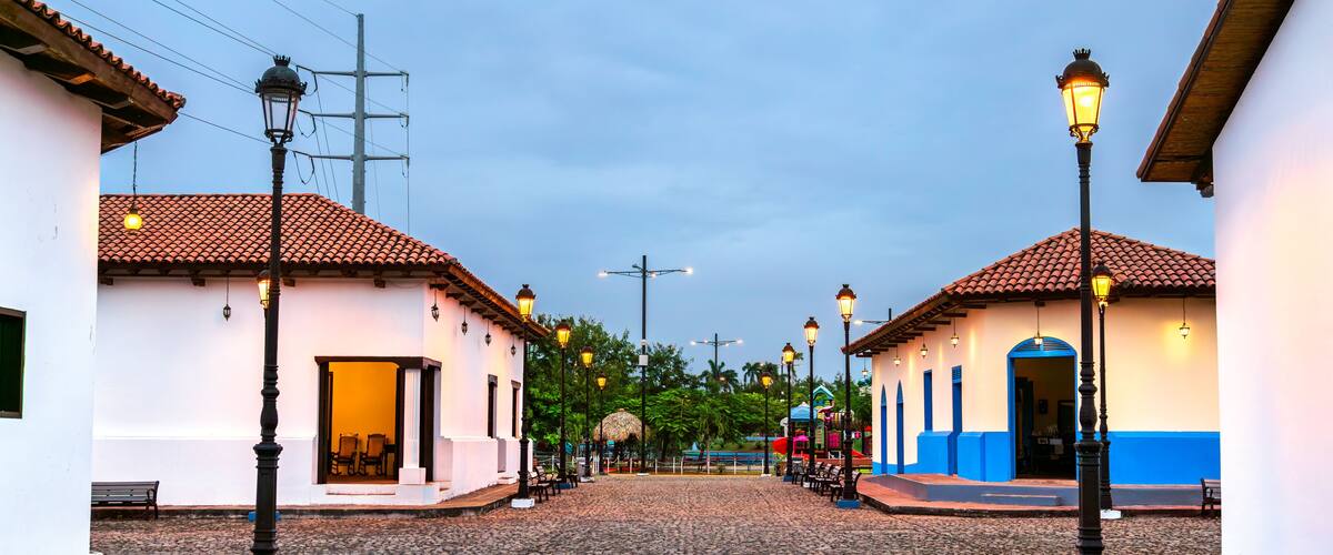 A cobblestone street with buildings, street lamps, and a thatched-roof structure.