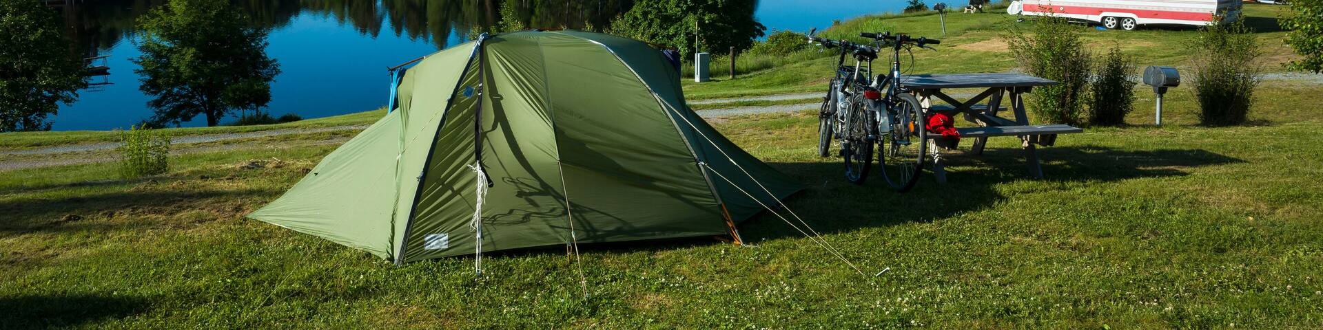 Green tent on a campsite grass, Bicycles stand next to a wooden picnic table, Sunny day blue sky, lake Ärtingen, Bengtsfors, Sweden