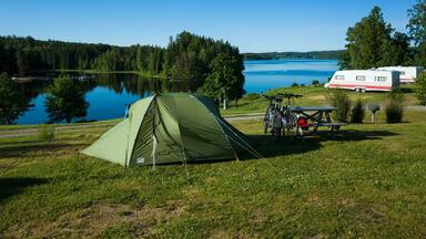 Green tent on a campsite grass, Bicycles stand next to a wooden picnic table, Sunny day blue sky, lake Ărtingen, Bengtsfors, Sweden