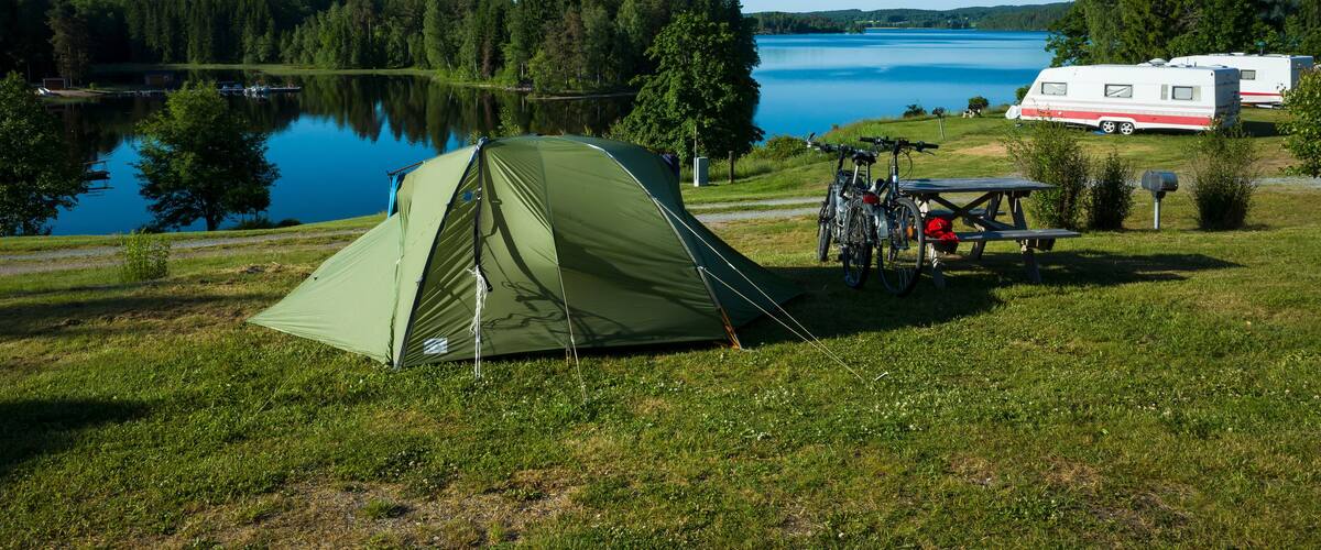 A green tent, a bicycle, a picnic table, and a white RV by a lake.