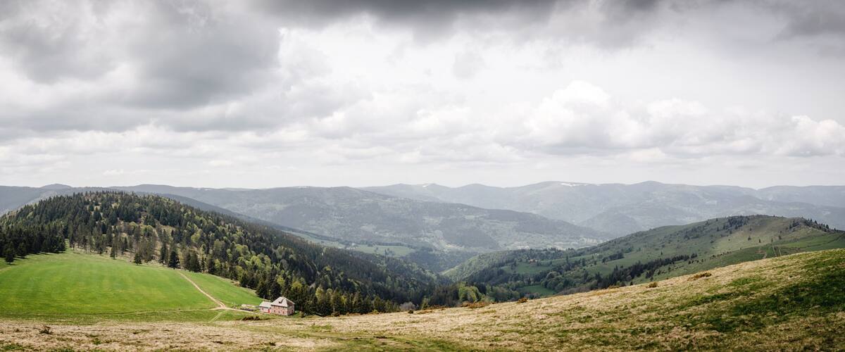 A rural landscape with rolling hills, a red house, and a forested mountain range under a cloudy sky.