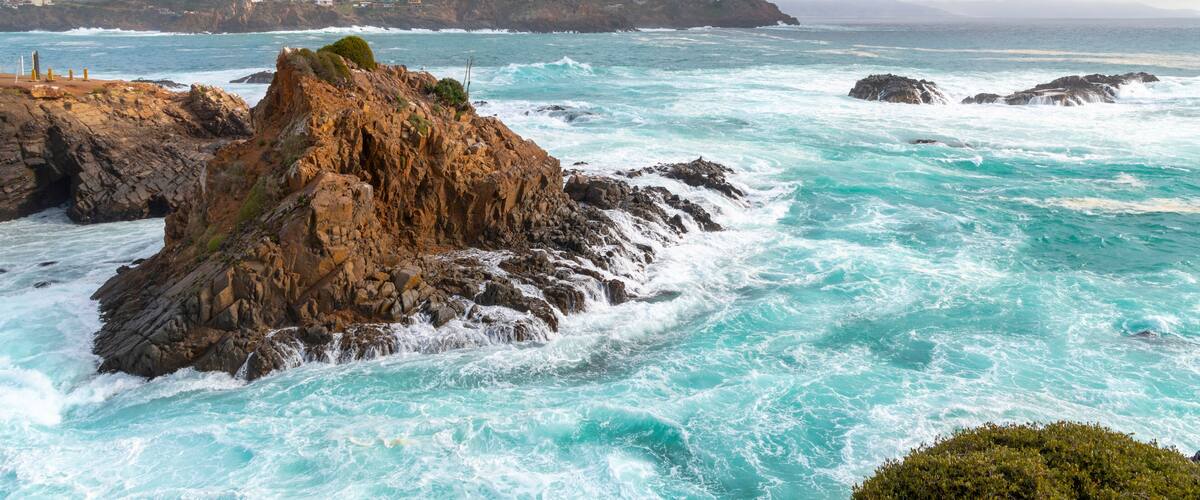 A rocky coastline with waves crashing against the rocks, a distant mountain range, and a cloudy sky.