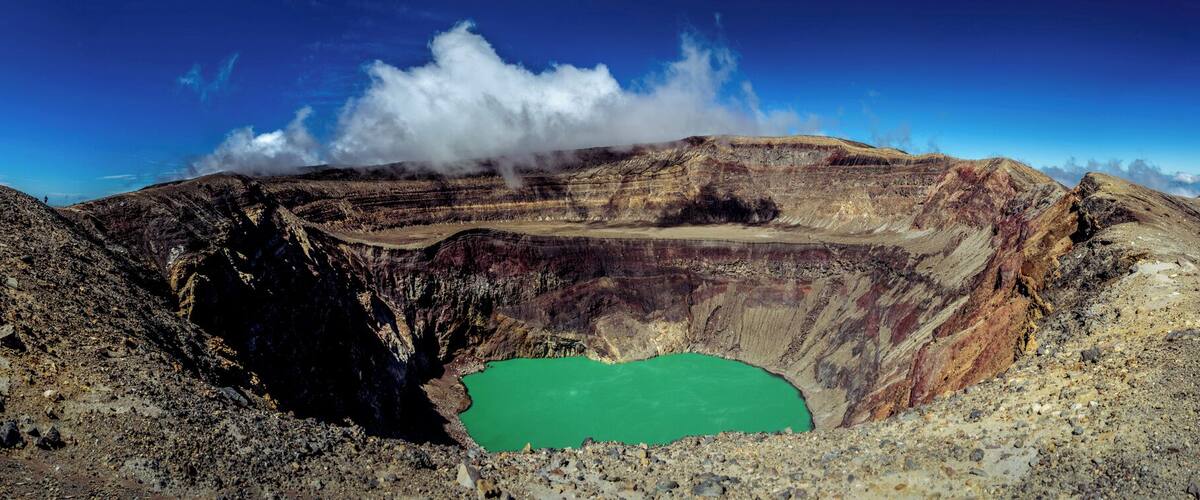 A volcanic crater with a green lake surrounded by rugged terrain and a group of people standing on a ridge.