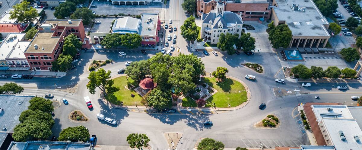 An aerial view of a city intersection with a central roundabout, surrounding buildings, and greenery.