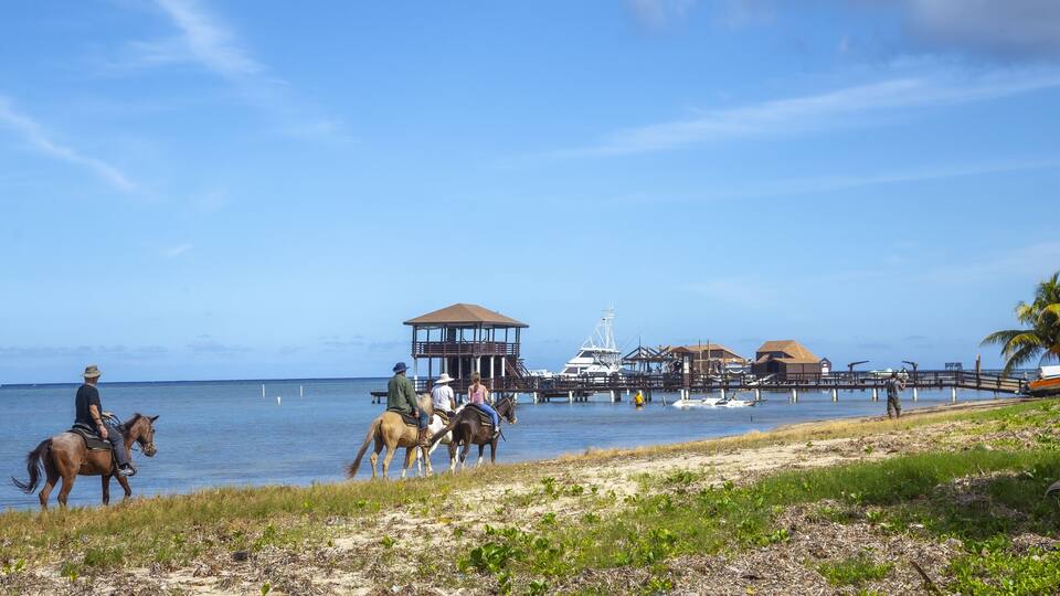 A beach with people riding horses, a pier, and a boat in the background.