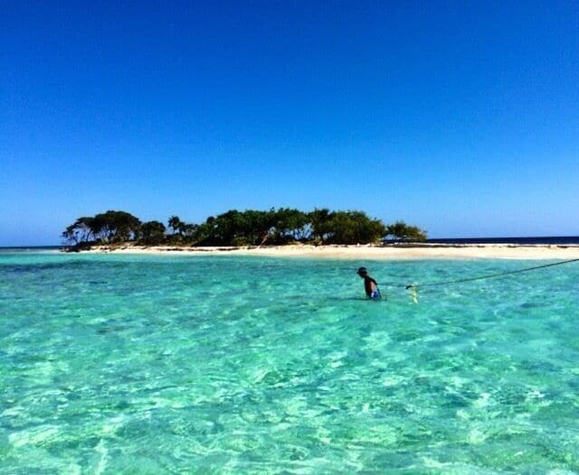 Água turquesa cristalina, uma praia arenosa e uma pequena ilha com árvores.