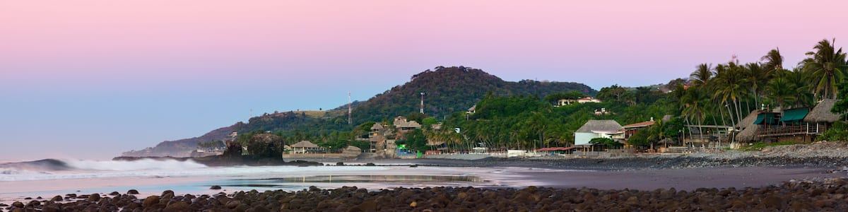 Eine Küstenlandschaft mit einem felsigen Strand, Wellen, die auf die Küste branden, einem Hügel mit Gebäuden und Palmen sowie einem rosafarbenen Himmel.