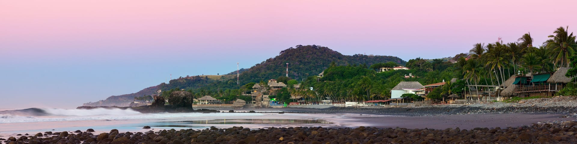 A coastal landscape with a rocky beach, waves crashing onto the shore, a hill with buildings and palm trees, and a pink sky.