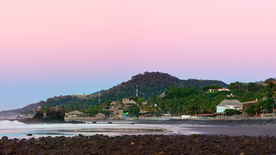 A coastal landscape with a rocky beach, waves crashing onto the shore, a hill with buildings and palm trees, and a pink sky.