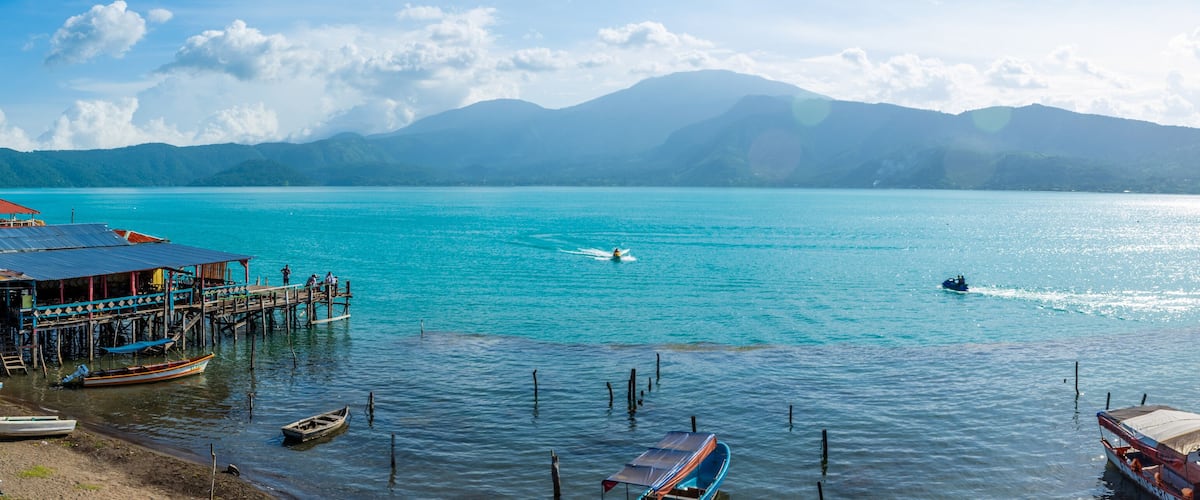 A turquoise lake with boats, a pier, and mountains in the background.