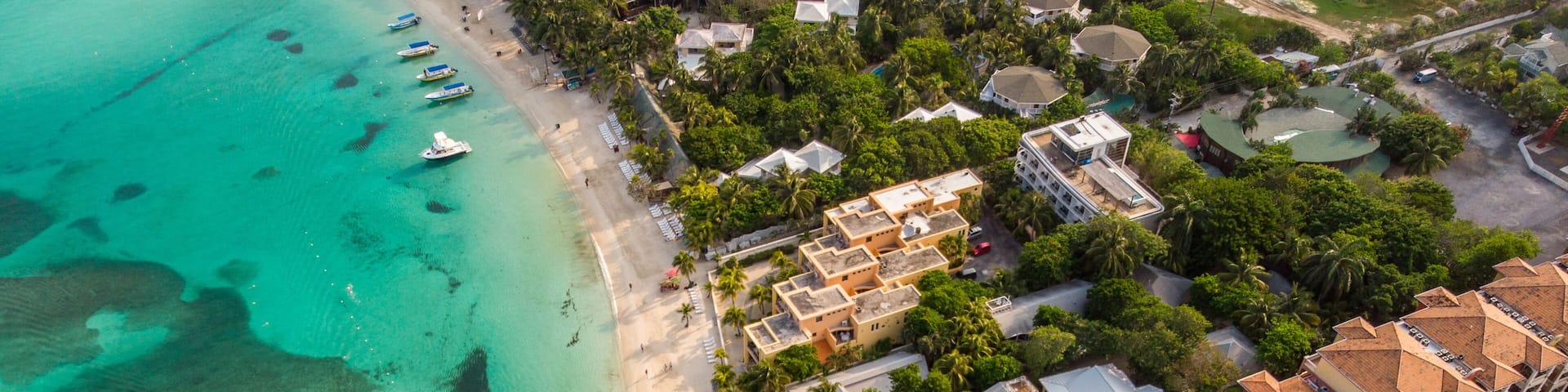 An aerial view of a resort with a beach, clear turquoise waters, a swimming pool, and resort buildings with red-tiled roofs.
