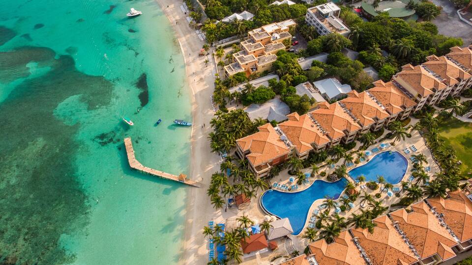 An aerial view of a resort with a beach, clear turquoise waters, a swimming pool, and resort buildings with red-tiled roofs.