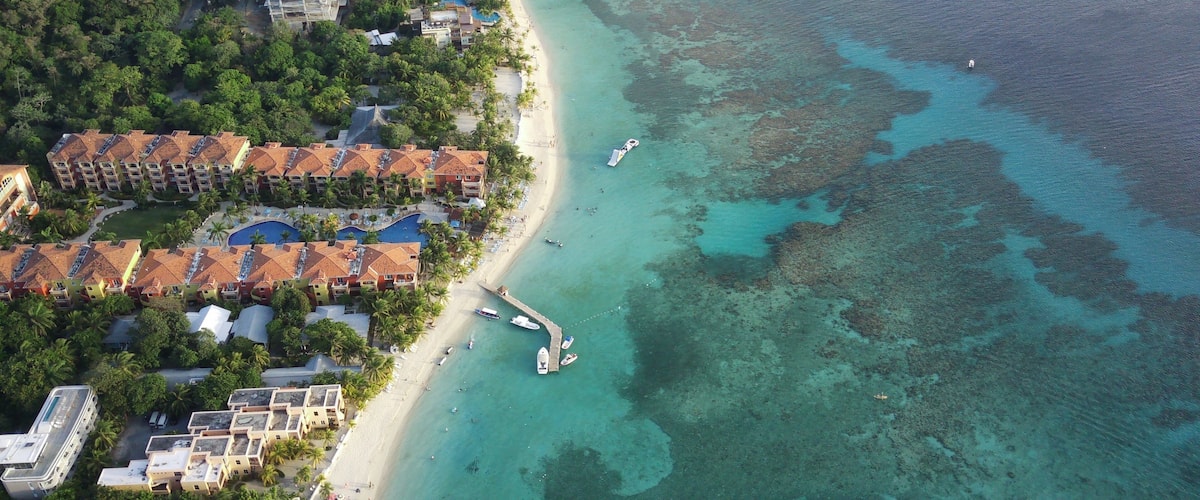 An aerial view of a coastal resort with a beach, clear turquoise waters, and a small pier.