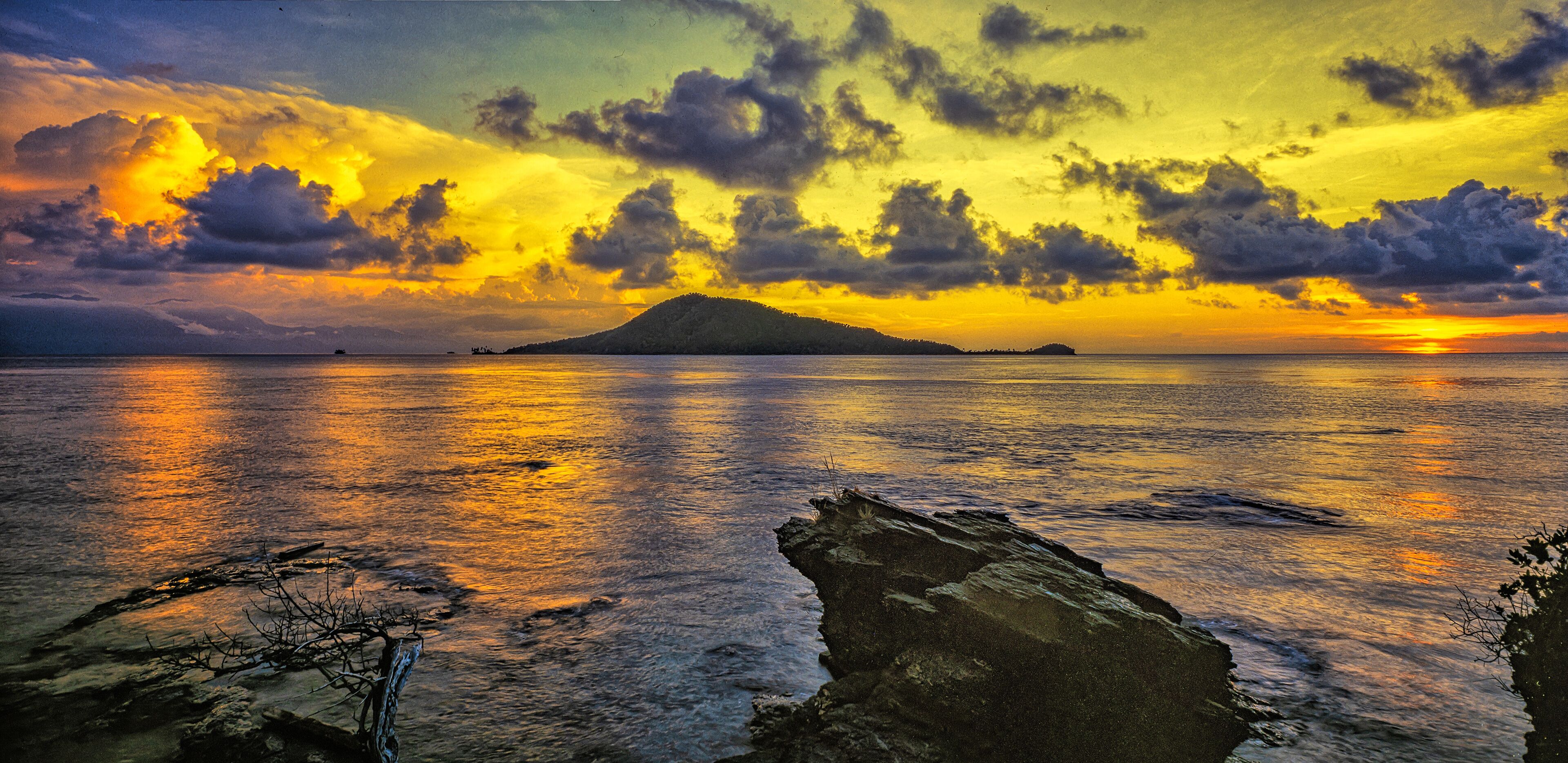 A sunset over a calm sea with a rocky foreground and an island in the distance.