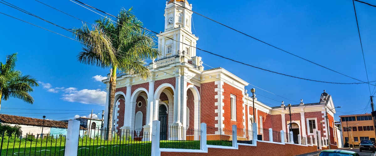 A historic building with a clock tower, surrounded by palm trees and a street with a vintage car.