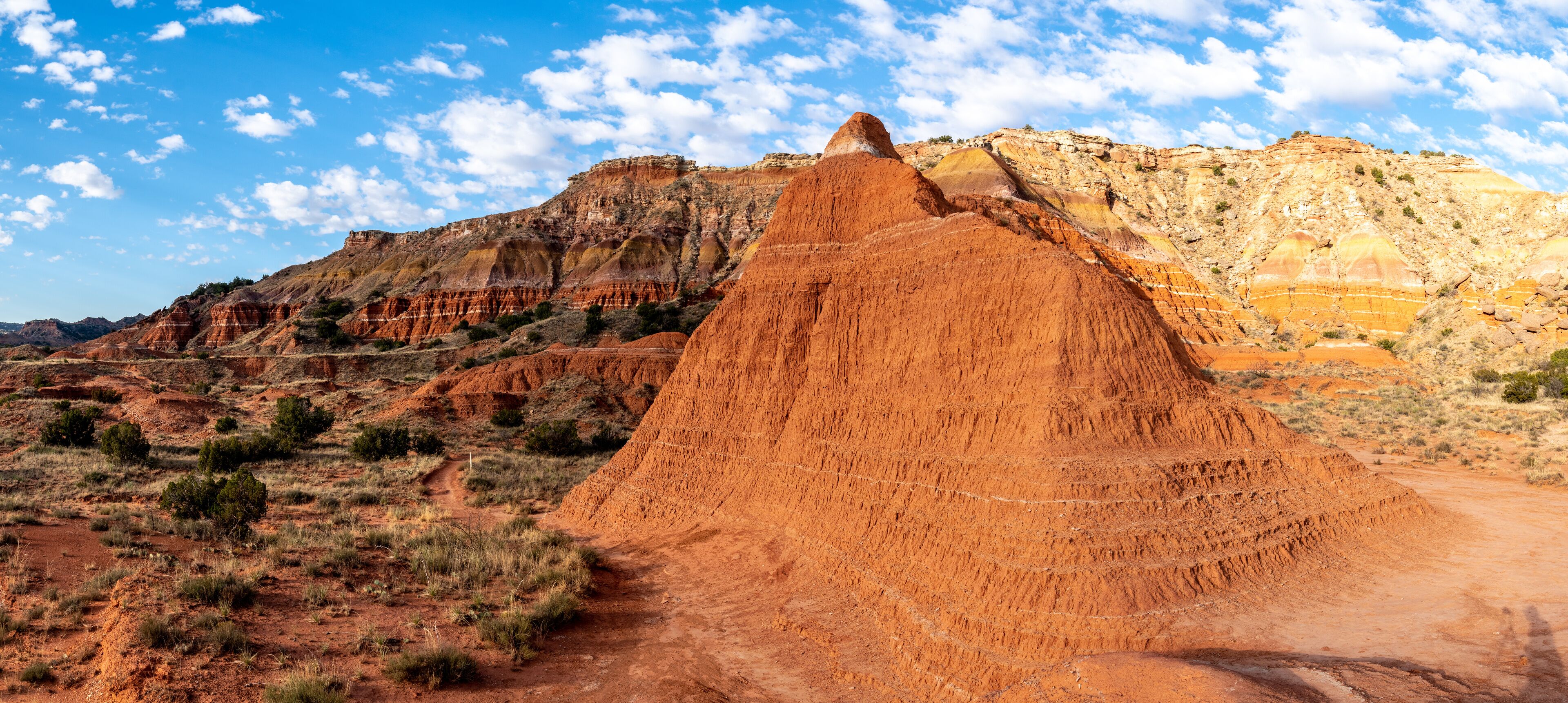 A mud cliff on a side of a mountain with striations of white, brown and yellow in the background rock and shrubs and grass and a hiking trail, Givens Spicer Lowery Trail, Palo Duro State Park, Texas