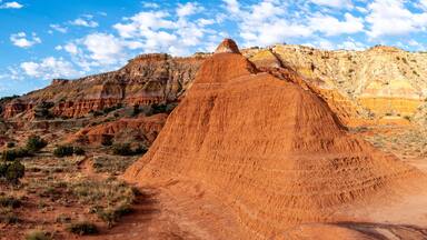 A mud cliff on a side of a mountain with striations of white, brown and yellow in the background rock and shrubs and grass and a hiking trail, Givens Spicer Lowery Trail, Palo Duro State Park, Texas