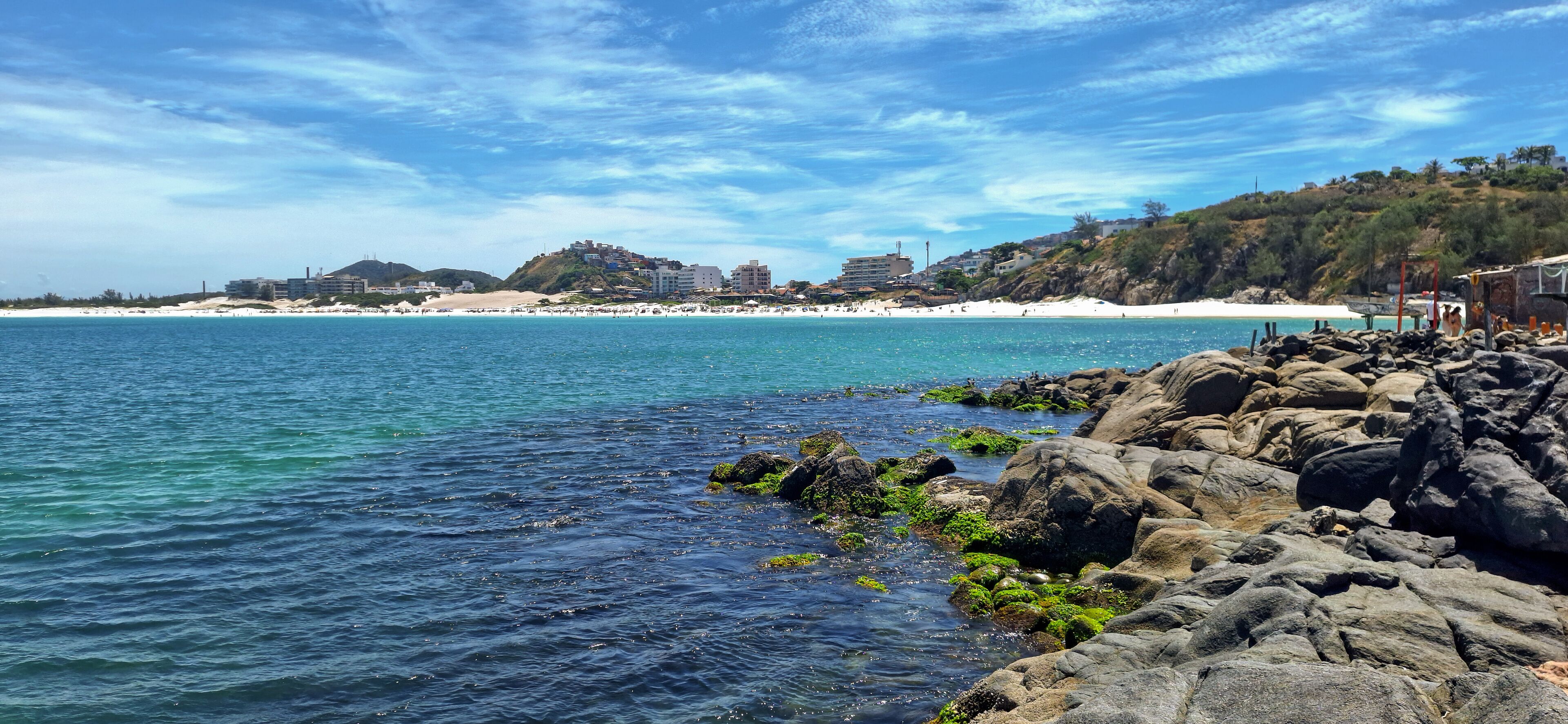 A coastal scene with clear blue waters, rocky shoreline, and a distant beach with buildings.