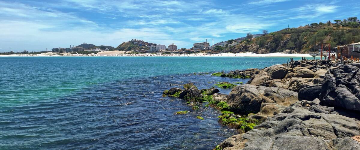A coastal scene with clear blue waters, rocky shoreline, and a distant beach with buildings.