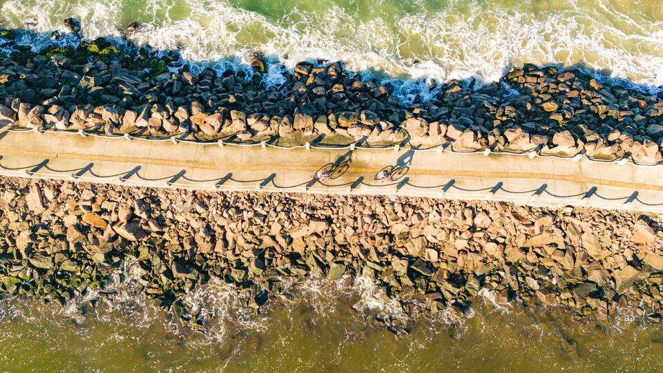 A coastal area with waves crashing onto a rocky shore, a concrete barrier, and a sandy beach.
