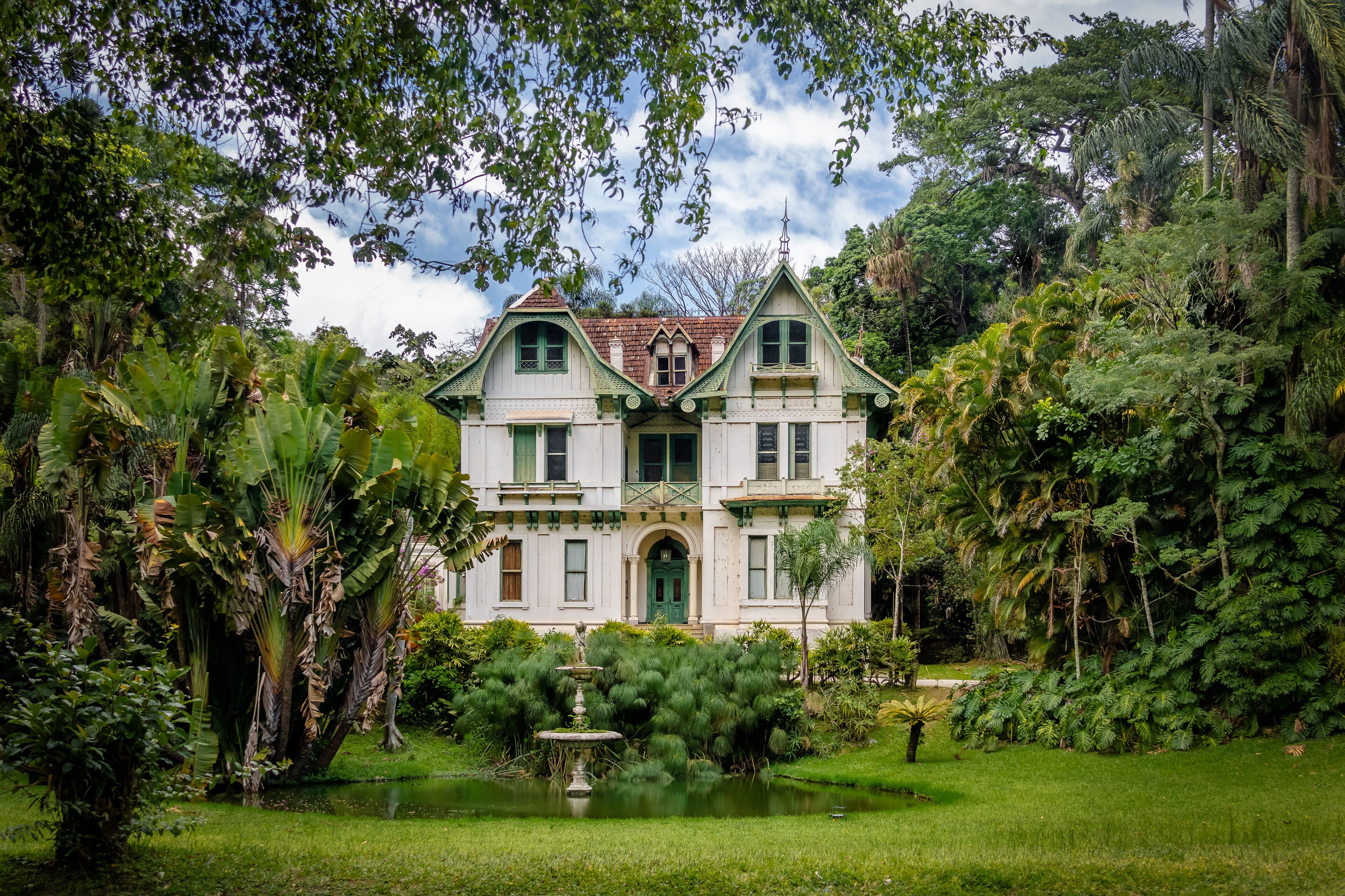 A large, white colonial-style house surrounded by lush tropical vegetation and a small pond.