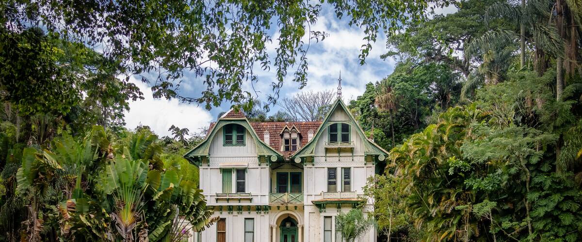A large, white colonial-style house surrounded by lush tropical vegetation and a small pond.