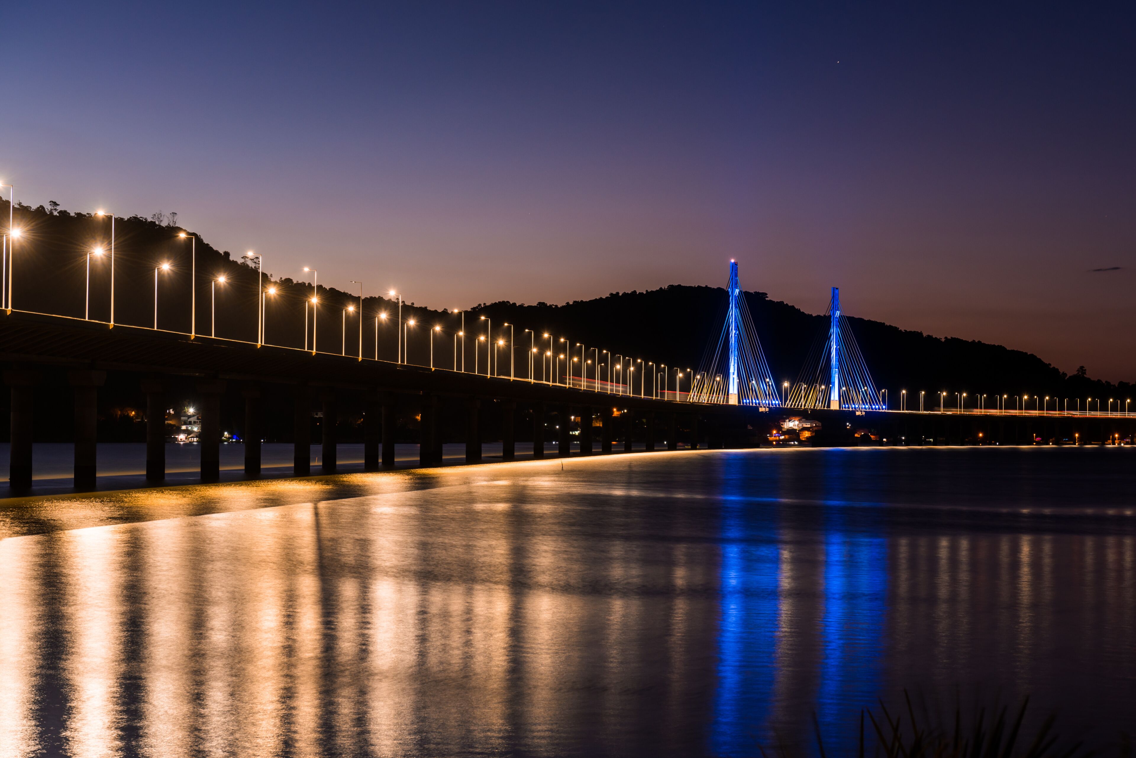 A nighttime view of a bridge with illuminated lights and a calm body of water reflecting the lights.