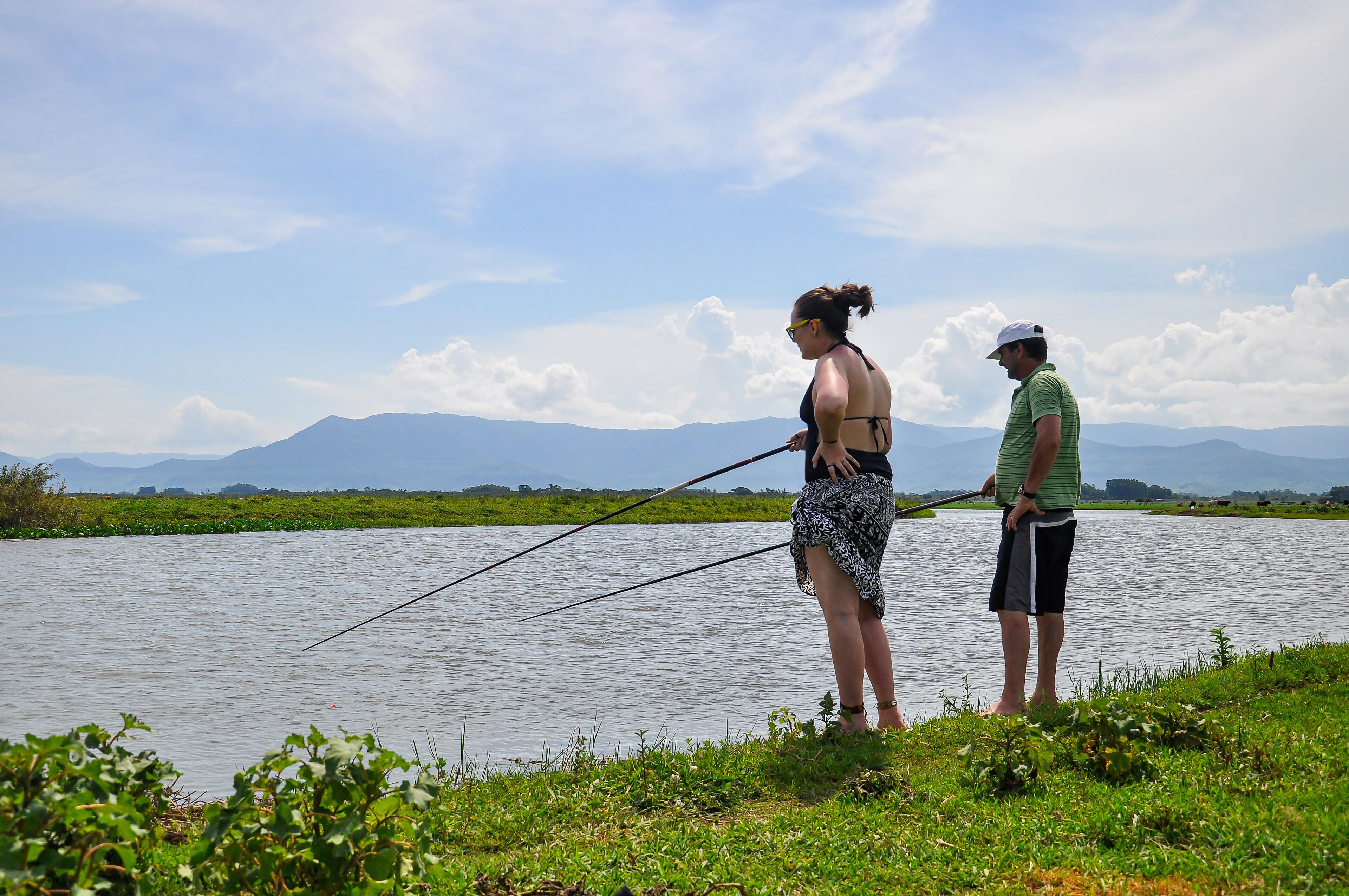 Two people fishing by a calm river with mountains in the background.