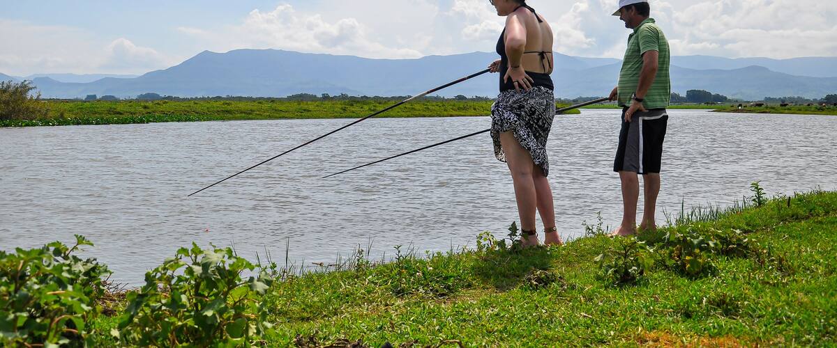 Two people fishing by a calm river with mountains in the background.