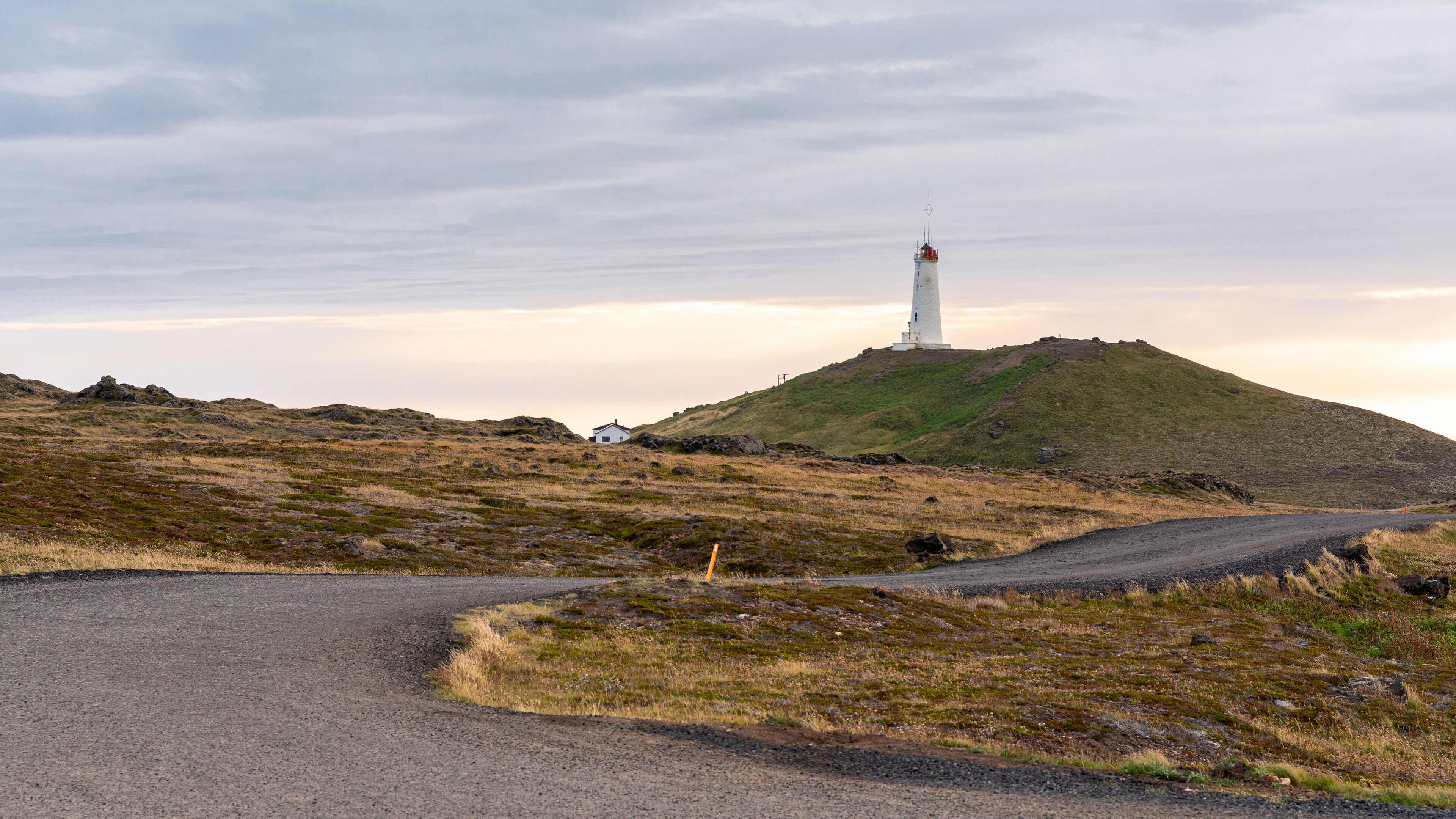 A white lighthouse on a grassy hill with a gravel road curving around it.