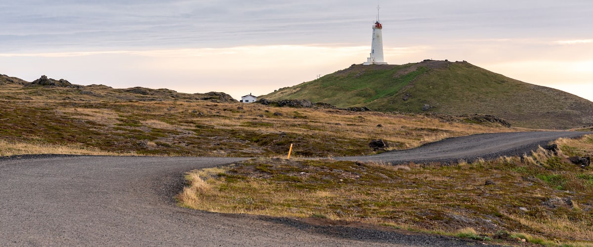A white lighthouse on a grassy hill with a gravel road curving around it.