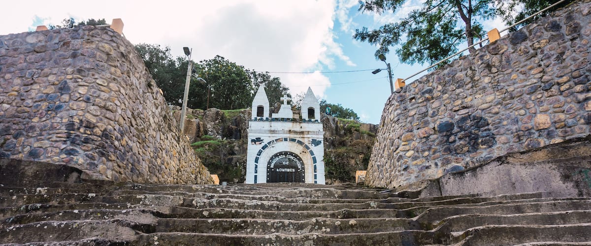 A stone staircase leading to a white church with a cross on top, surrounded by stone walls and trees.